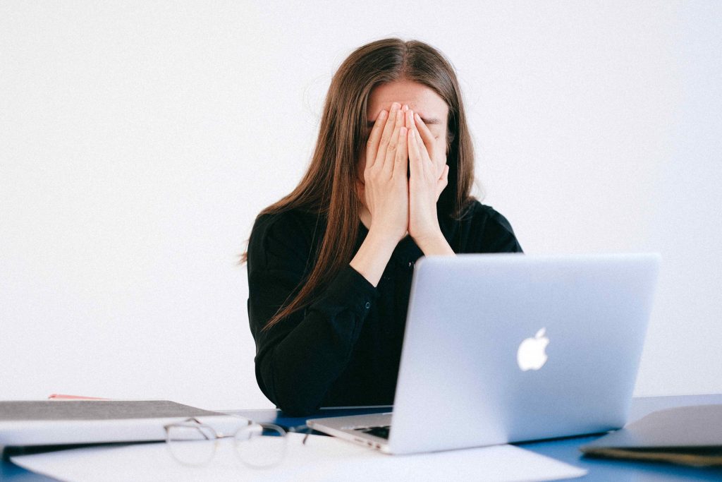 Cropped image of brown haired woman sitting a desk in front of laptop holding her face in her hands experiencing the symptoms of peri and menopause.