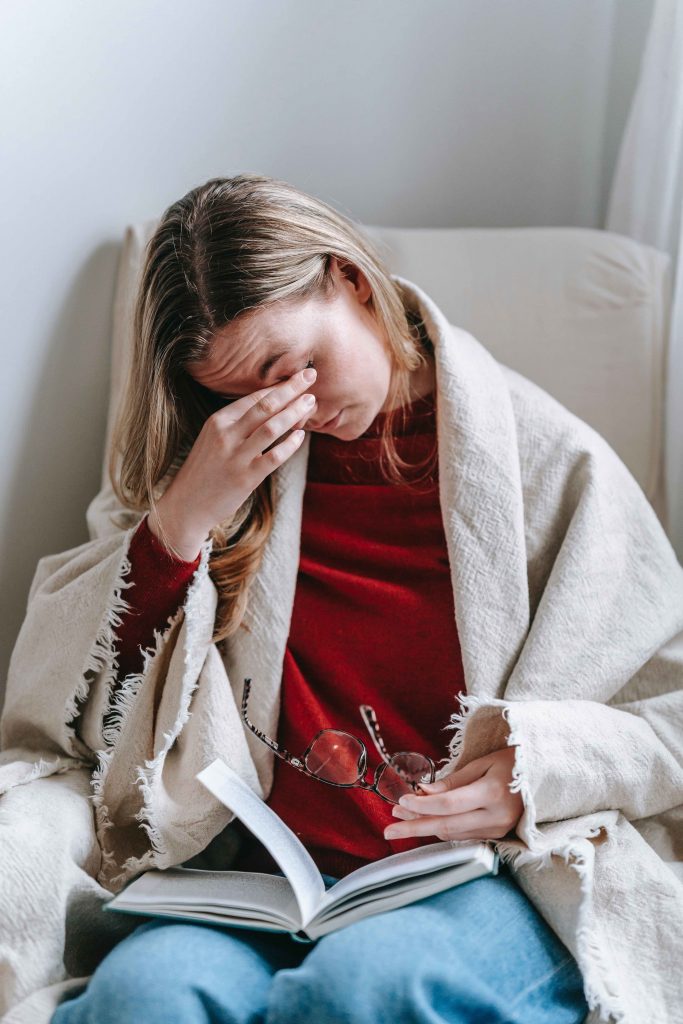 Cropped image of woman with blonde hair sitting on armchair with book in lap rubbing eyes experiencing symptoms of peri and menopause.