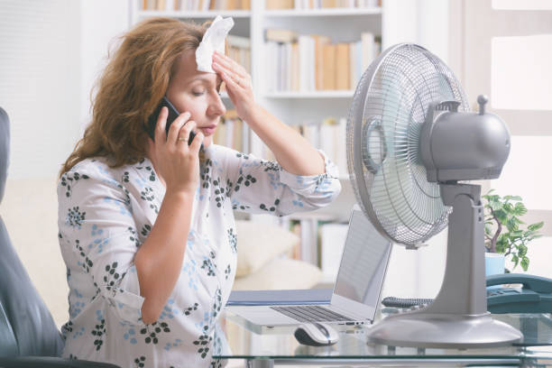 Cropped image of woman sitting at a desk in front of pedestal fan wiping brow of sweat from the symptoms of peri and menopause.
