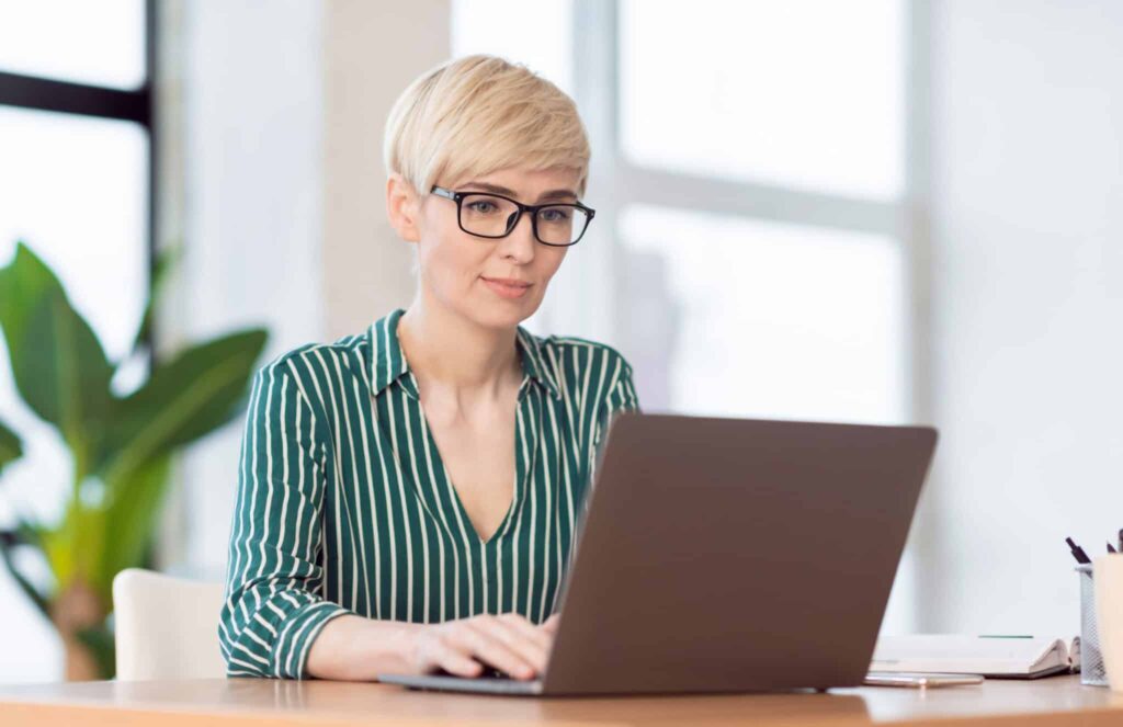 Cropped image of woman with short blonde hair wearing a green and white striped top using a laptop completing her registering her work colleagues in the Don't Sweat It Peri and Menopause online course.