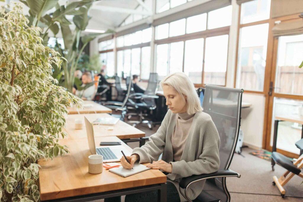 Cropped image of blonde hair woman sitting at timber desk in a share office taking notes on a notepad about how to navigate peri and menopause while maintaining career progression.