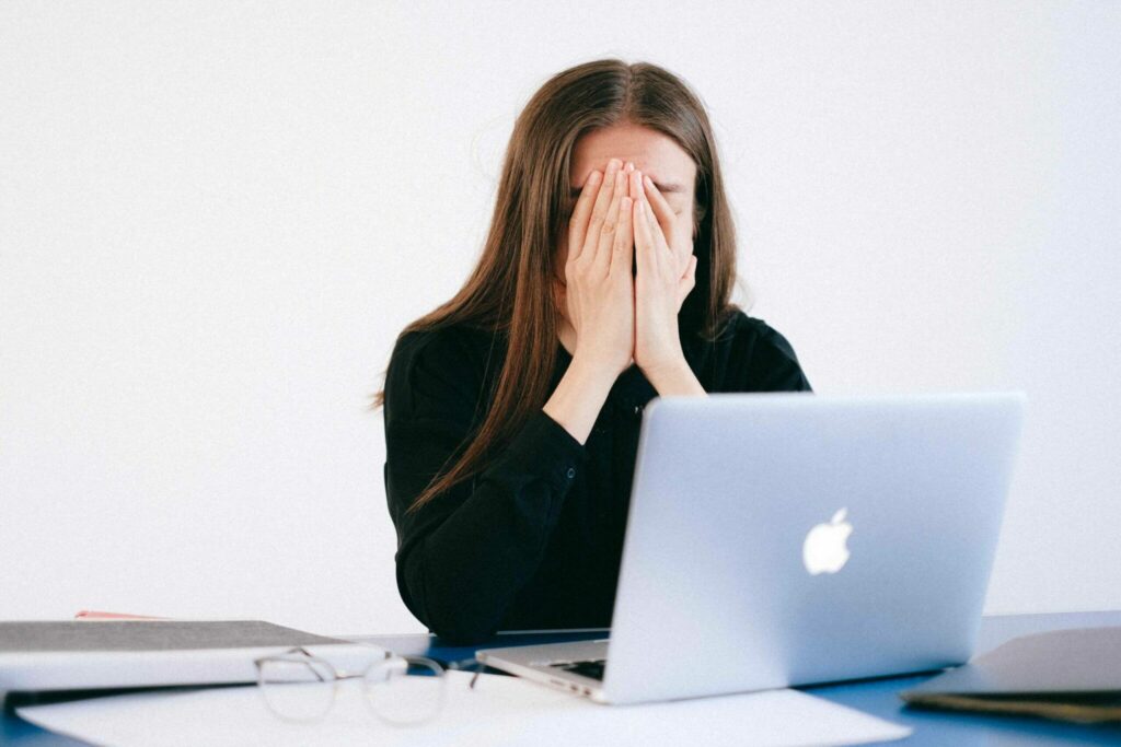Cropped image of brown haired woman sitting a desk in front of laptop holding her face in her hands experiencing the symptoms of peri and menopause.