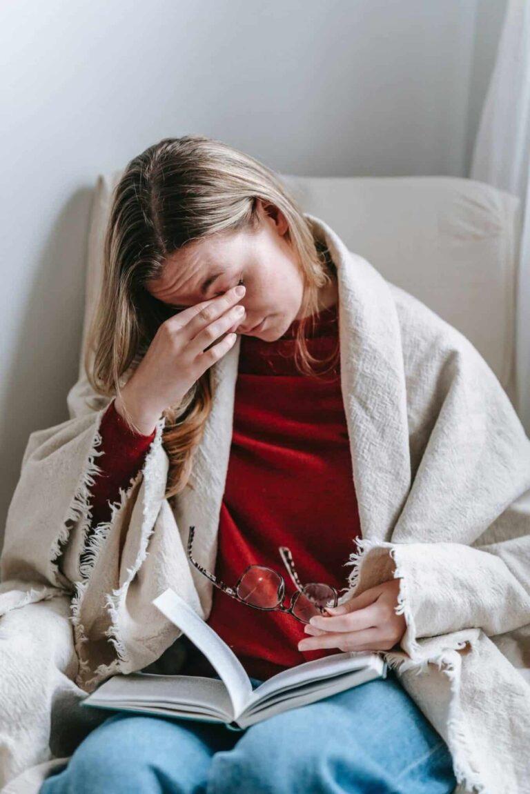 Cropped image of woman with blonde hair sitting on armchair with book in lap rubbing eyes experiencing symptoms of peri and menopause.